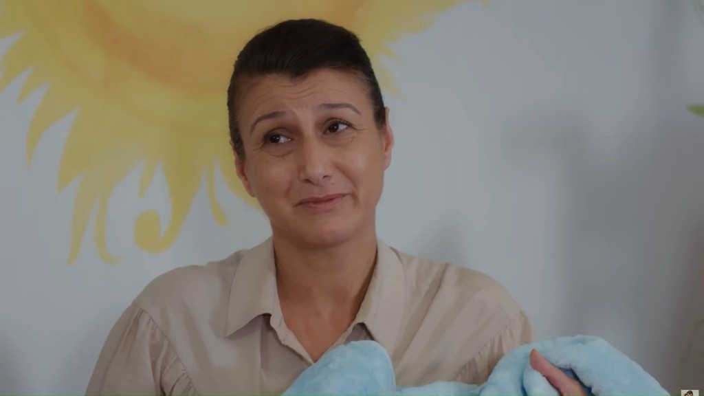 Zbliżenie na kobietę ze spiętymi włosami trzymającą zawinięte niemowlę; łagodny, uśmiechnięty wyraz twarzy, tło z malunkiem słońca.