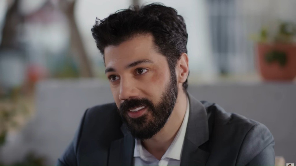 Melih przechyla głowę i uśmiecha się.