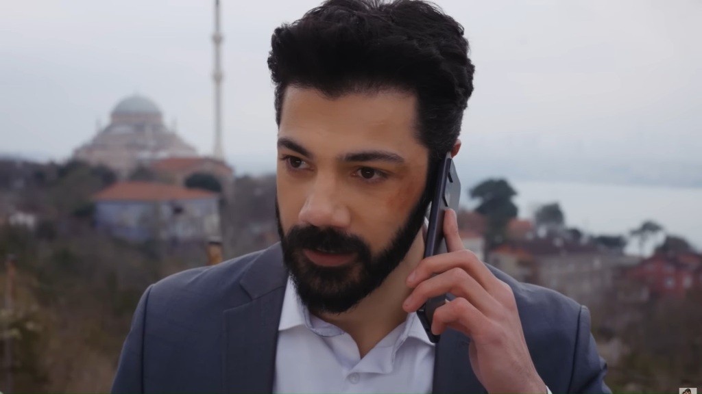 Zbliżenie na Meliha, który rozmawia przez telefon. W tle budynki oraz szare niebo.