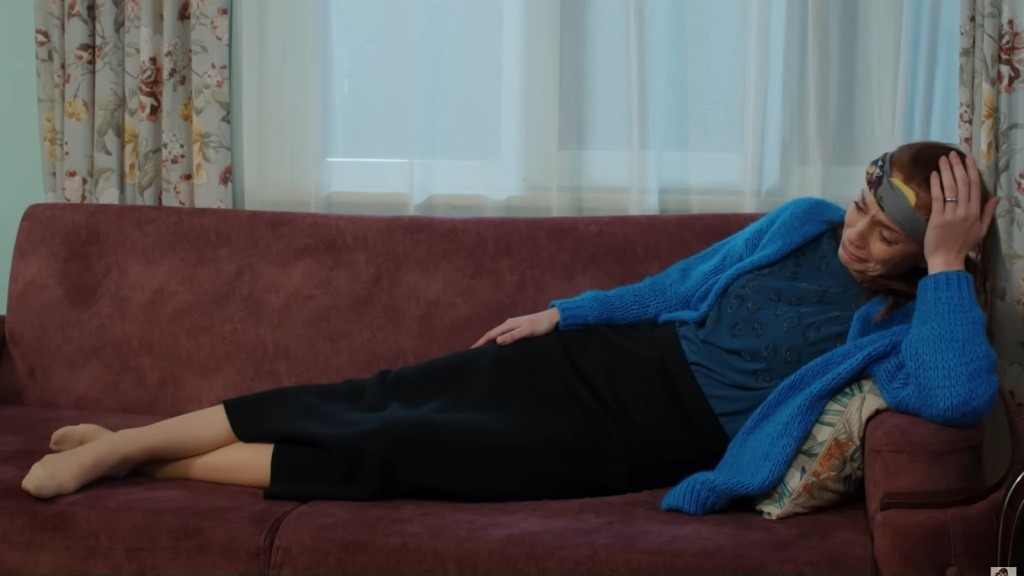 Derya w niebieskim kardiganie leży zamyślona na kanapie.