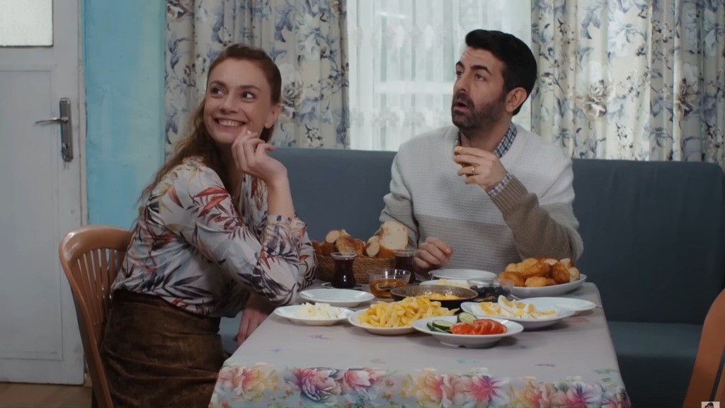 Derya i Cemil siedzą przy stole z przekąskami i filiżankami w domowym wnętrzu. Patrzą w jednym kierunku.