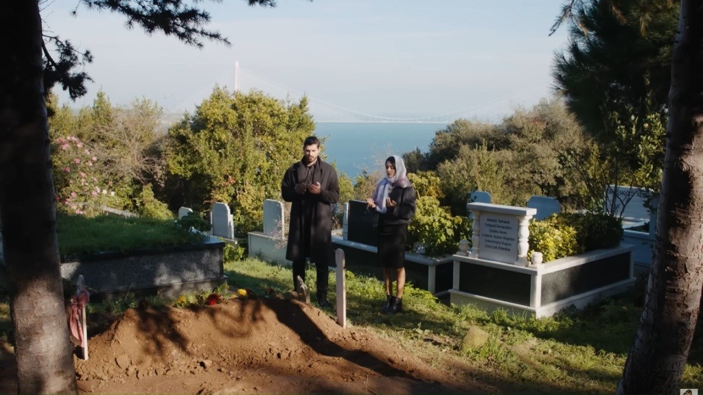 Cihan i Hancer stojący przy świeżo wykopanym grobie na zielonym wzgórzu z widokiem na morze — smutna, refleksyjna scena.