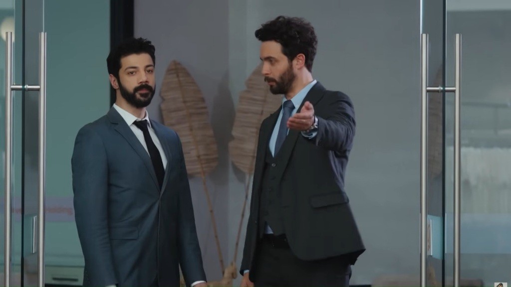 Engin wyciąga rękę przed siebie, zapraszając Meliha do środka. Obaj są ubrani w eleganckie garnitury.