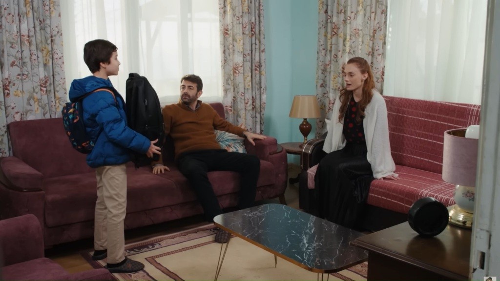 Cemil i Derya siedzą na kanapach. Emir stoi przed nimi, trzymając przed sobą duży czarny plecak.