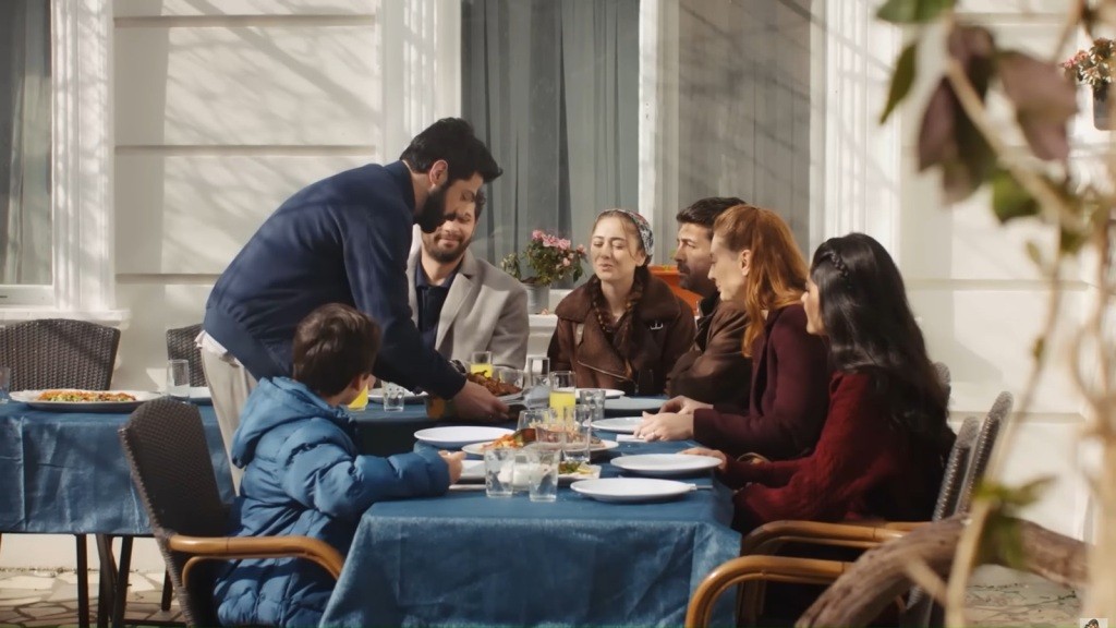 Melih kładzie talerz z grillowanym mięsem na stole przed starym domem. Przy stole siedzą Engin, Sila, Derya, Cemil, Hancer i Emir.