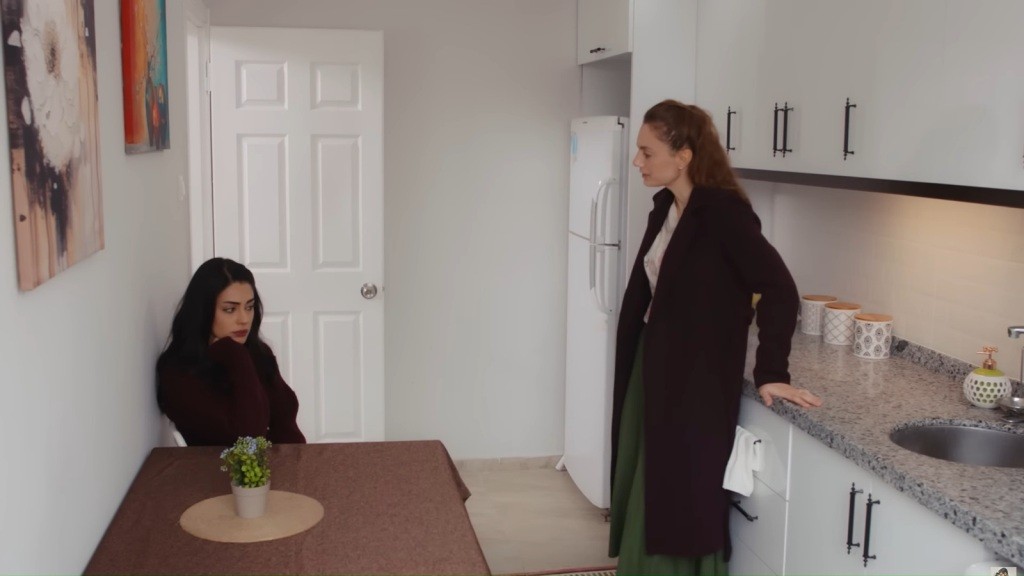 Hancer siedzi przy stole w kuchni. Derya opiera się o blat.
