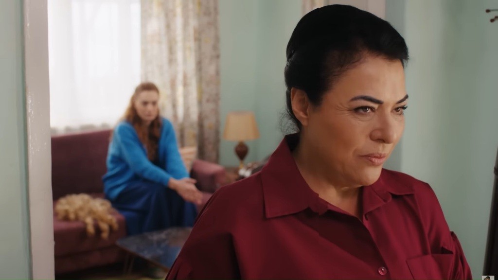 Mukadder uśmiecha się za progiem salonu, w którym siedzi Beyza na kanapie.