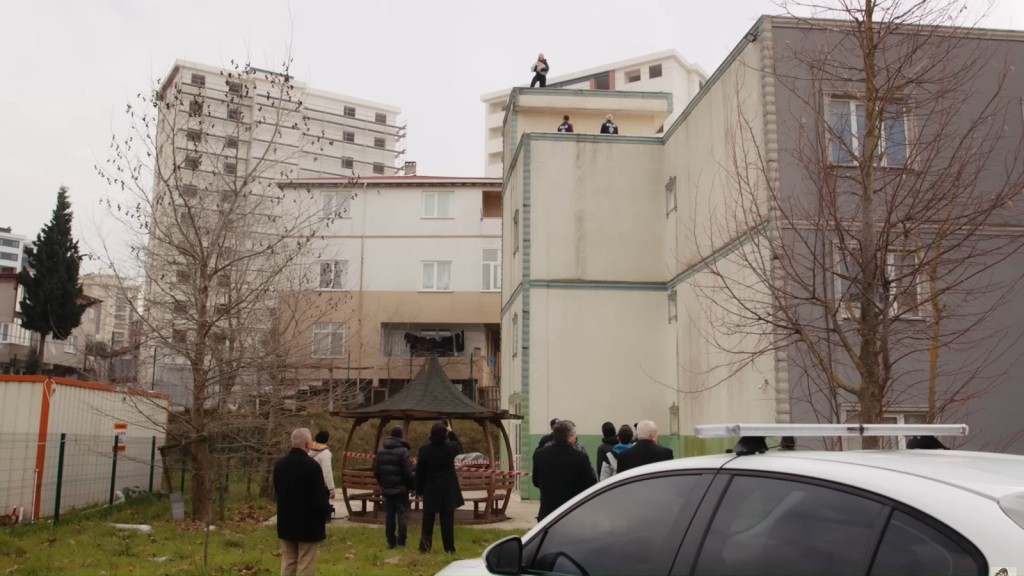 Widok na blok, na dachu którego stoi Yonca. Przed blokiem zgromadzeni są gapie.