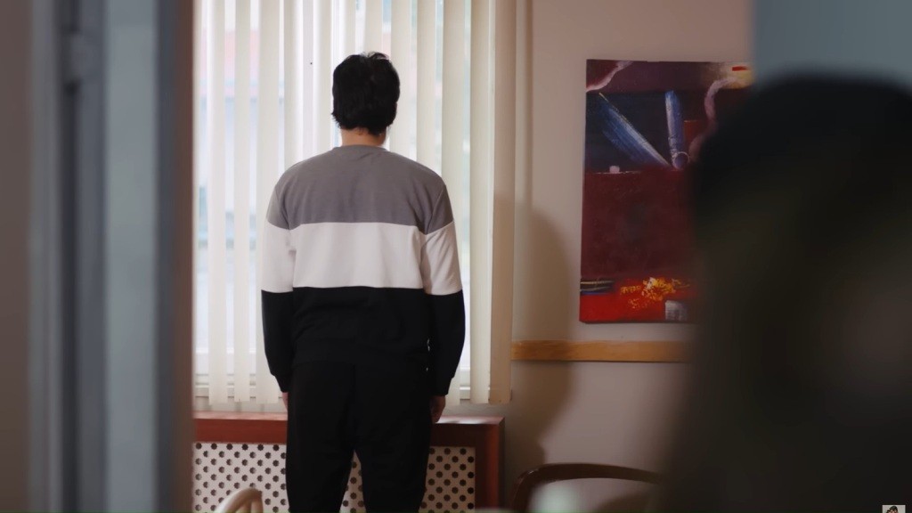 Widok do środka sali przez uchylone drzwi. Mężczyzna stoi nieruchomo przy oknie, odwrócony plecami.