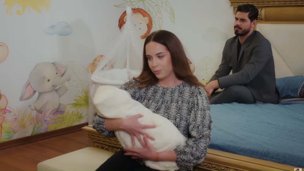 Beyza siedzi na ławce i trzyma dziecko na rękach. Cihan siedzi za nią na łóżku i patrzy na nią.