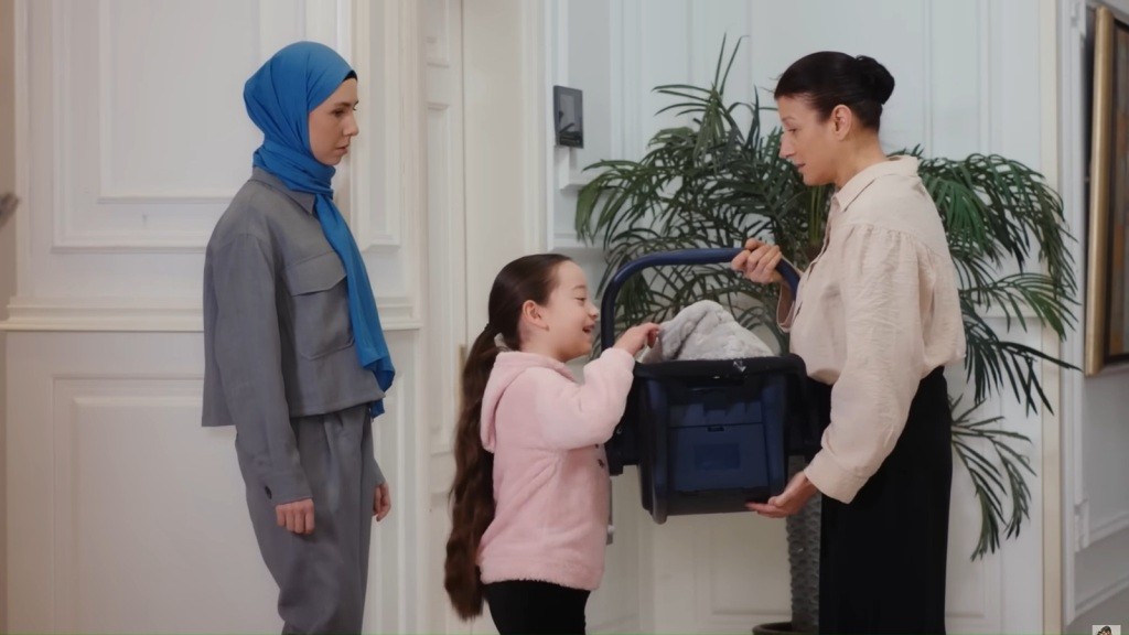 Mine zagląda do środka fotelika trzymanego przez Fadime. Tuż za dziewczynką stoi jej mama, Sinem.
