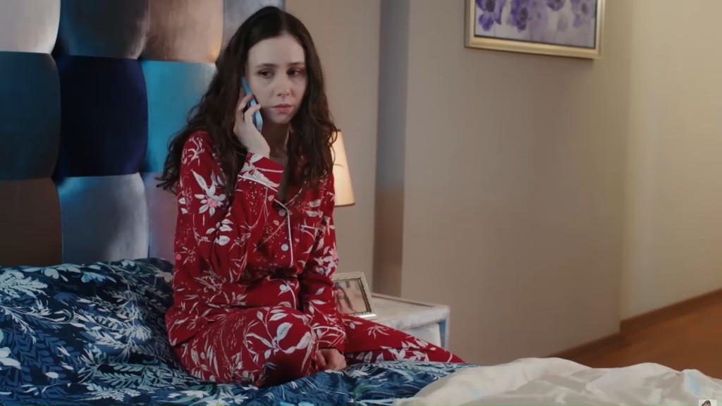 Sinem, ubrana w piżamę, siedzi na łóżku i rozmawia przez telefon.