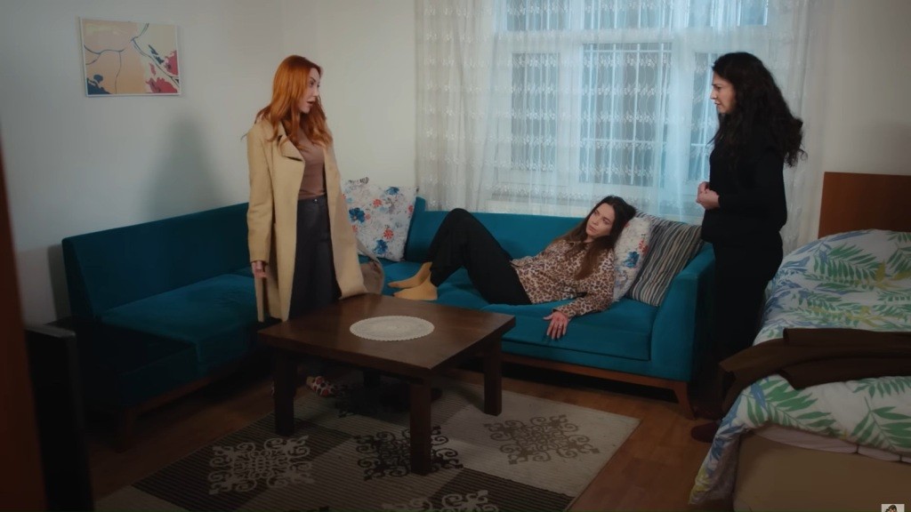 Beyza leży na turkusowej kanapie. Yonca i Gulsum stoją.
