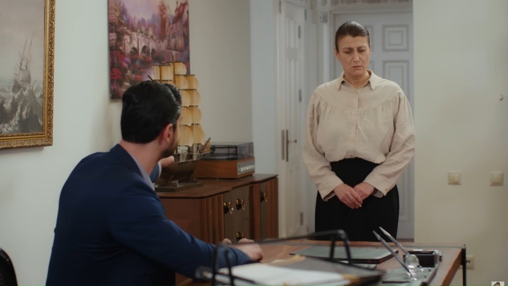 Fadime stoi przed biurkiem Cihana ze spuszczonym wzrokiem i splecionymi z przodu dłońmi.