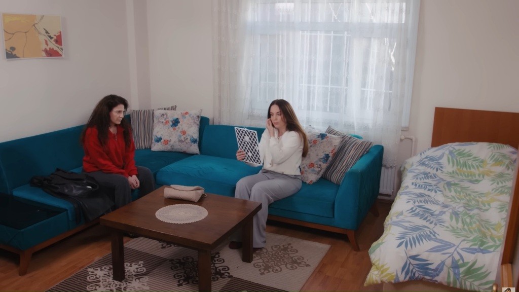 Gulsum i Beyza siedzą na turkusowej kanapie. Beyza trzyma w ręku lusterko i poprawia włosy.